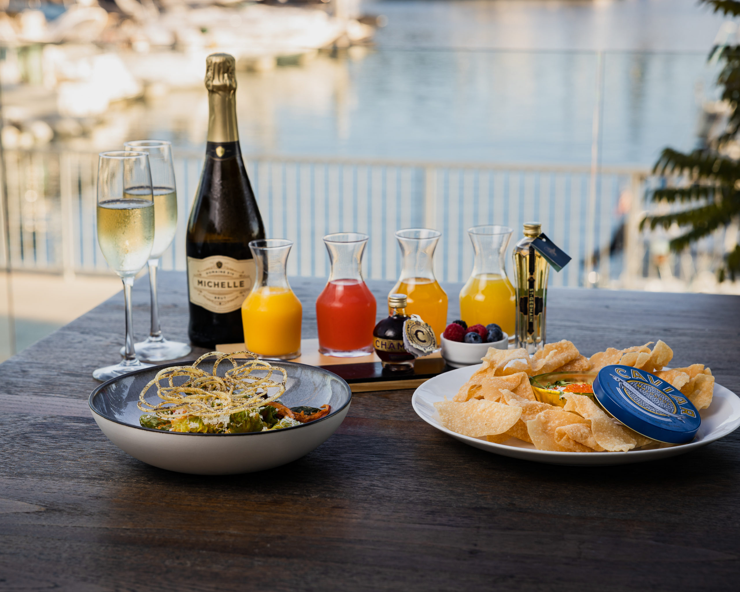mimosa drinks with two dishes of food and champagne bottle on table facing water and boats