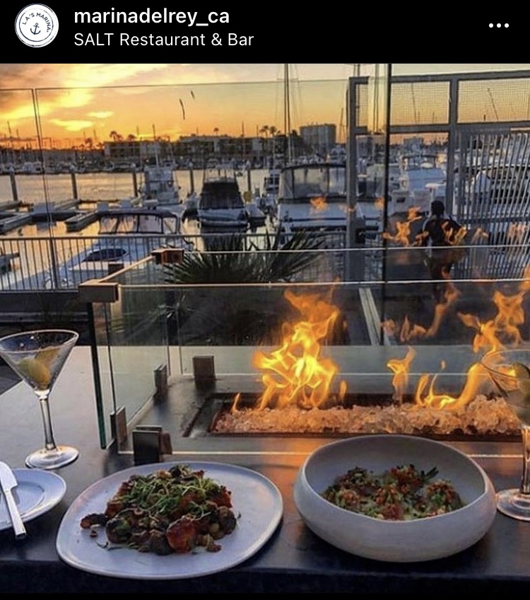 Dishes of food and cocktail glass by firepit table overlooking boats, sea and sunset