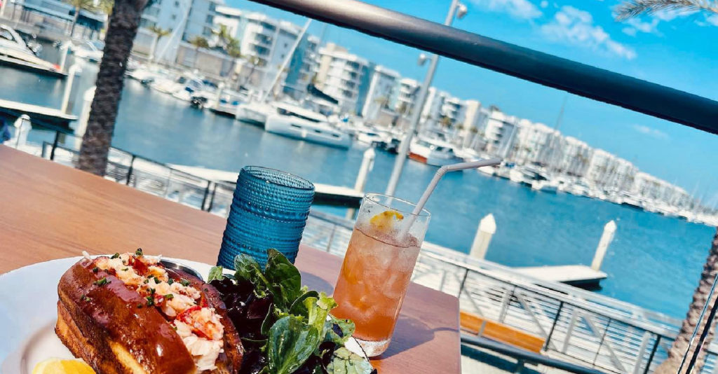 lobstser roll and salad on a dish with drinks outside facing harbor and ocean